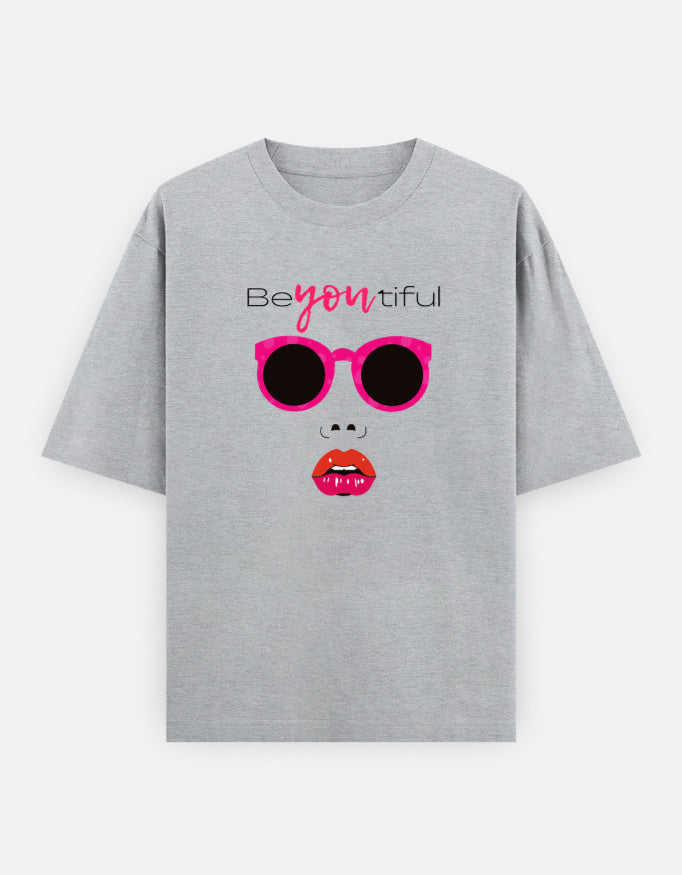 grey oversized unisex t shirt featuring a front print of a minimalist fashion face with bright pink sunglasses, bold pink lips, and the word Beyoutiful above.